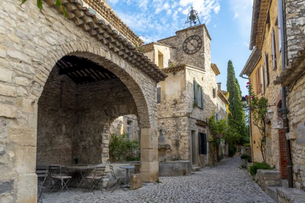 Rue pavée, ou calade, dans le centre du village, avec un proche qui abrite des tables et des chaises, une fontaine et clocher avec une horloge.