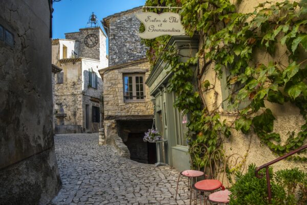 Rue pavée, ou calade, avec la devanture d'un salon de thé à droite et un clocher avec pendule et campanile.