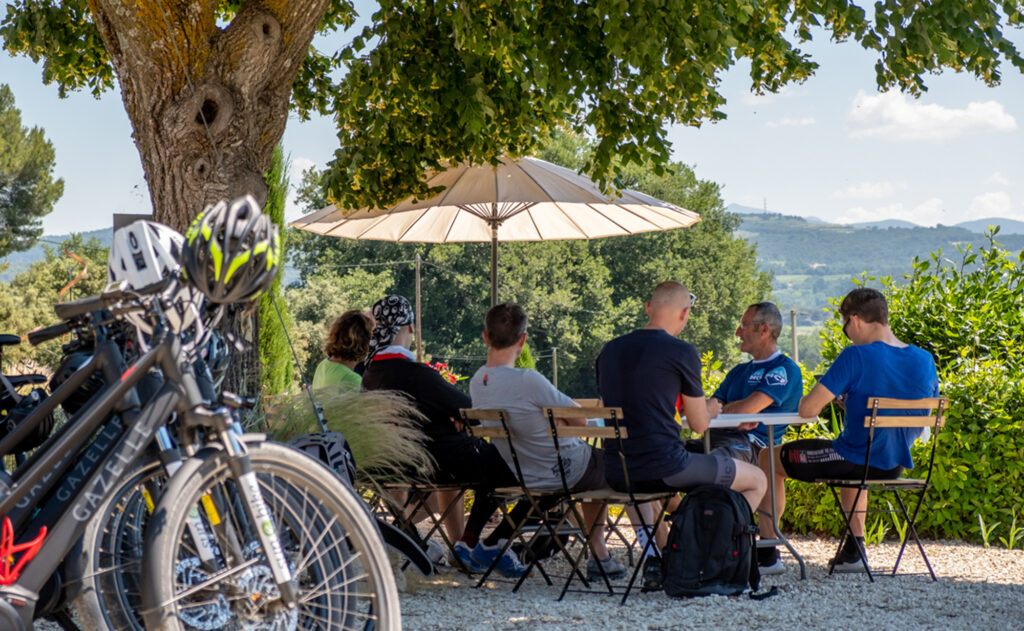 Un groupe de cyclistes installé à la terrasse d'un café, à l'ombre d'un parasol pour une pause fraîcheur.